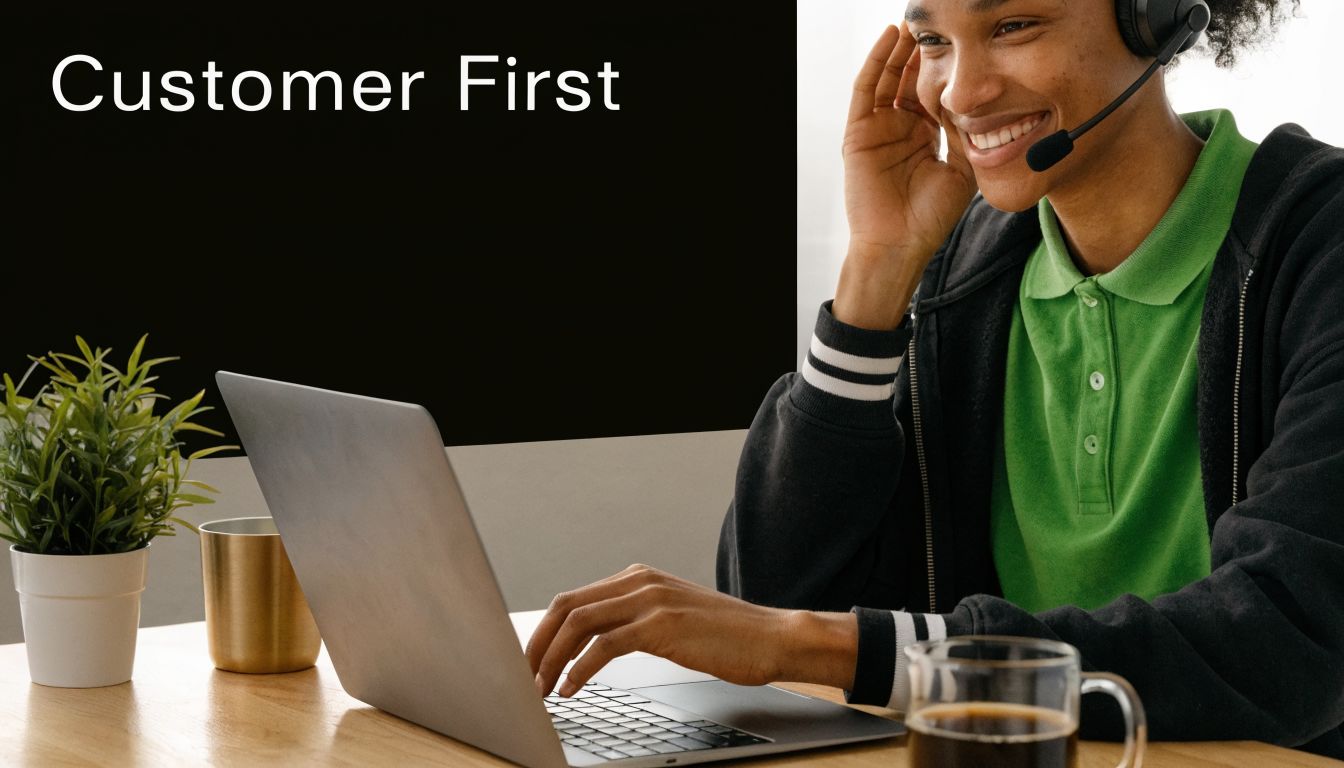 A smiling customer service representative using a headset and laptop at a desk with coffee and plants.